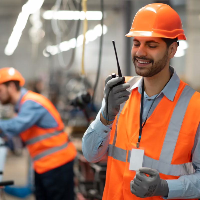 Industrial worker wearing an orange hard hat, high-visibility safety vest, and gloves communicates using a handheld two-way radio inside a modern manufacturing facility, while another worker operates equipment in the background. Bright overhead lighting and industrial machinery create an active workplace environment.